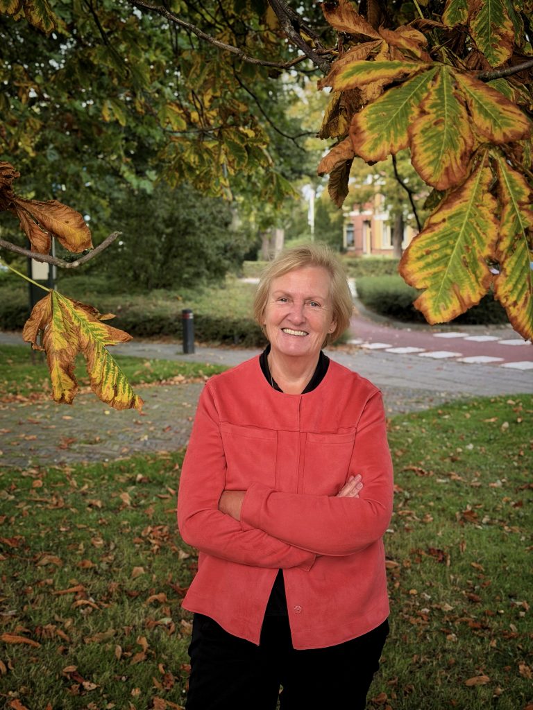 Wethouder Mary van Gent poseert in een rood jasje met armen over elkaar onder een boom tijdens de herfst