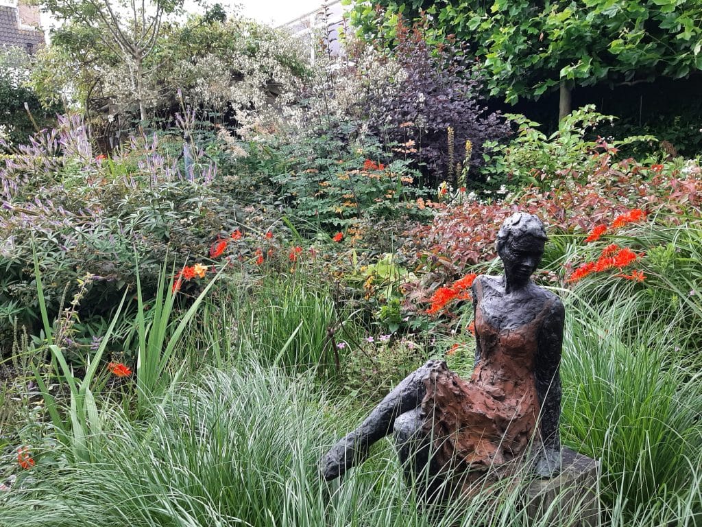 Beeld van een zittende figuur tussen bloemen en siergrassen in een groene tuin