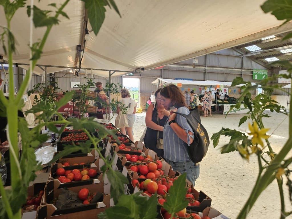 “Bezoekers bekijken tomaten en andere verse producten bij een marktkraam in een overdekte markthal, met planten op de voorgrond.”