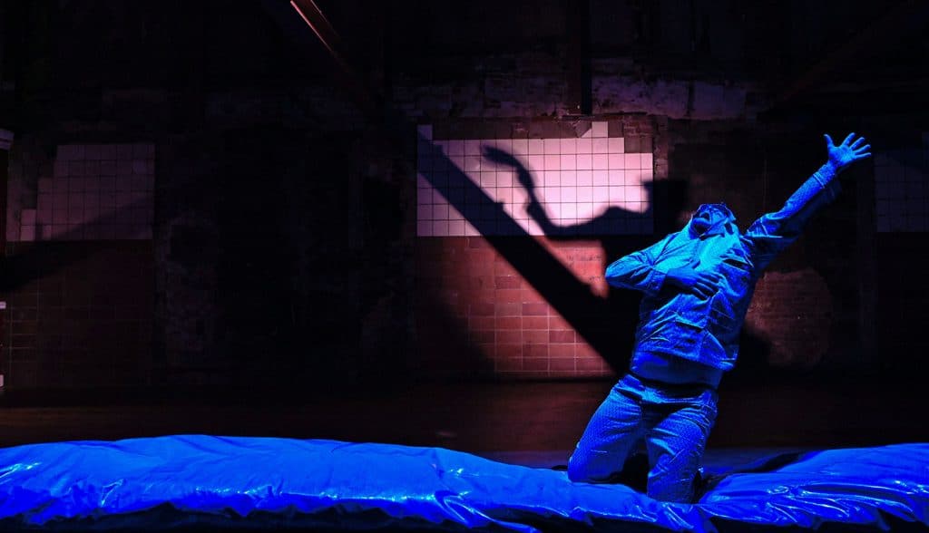 Theaterscène met performer in blauw licht, knielend op een podium met sterke schaduwen op de achtergrond.