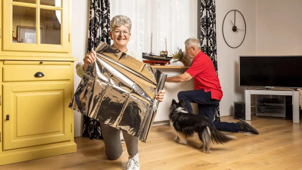 Een oudere vrouw toont isolatiefolie glimlachend aan de camera, terwijl een man op de achtergrond de folie aanbrengt achter een radiator.