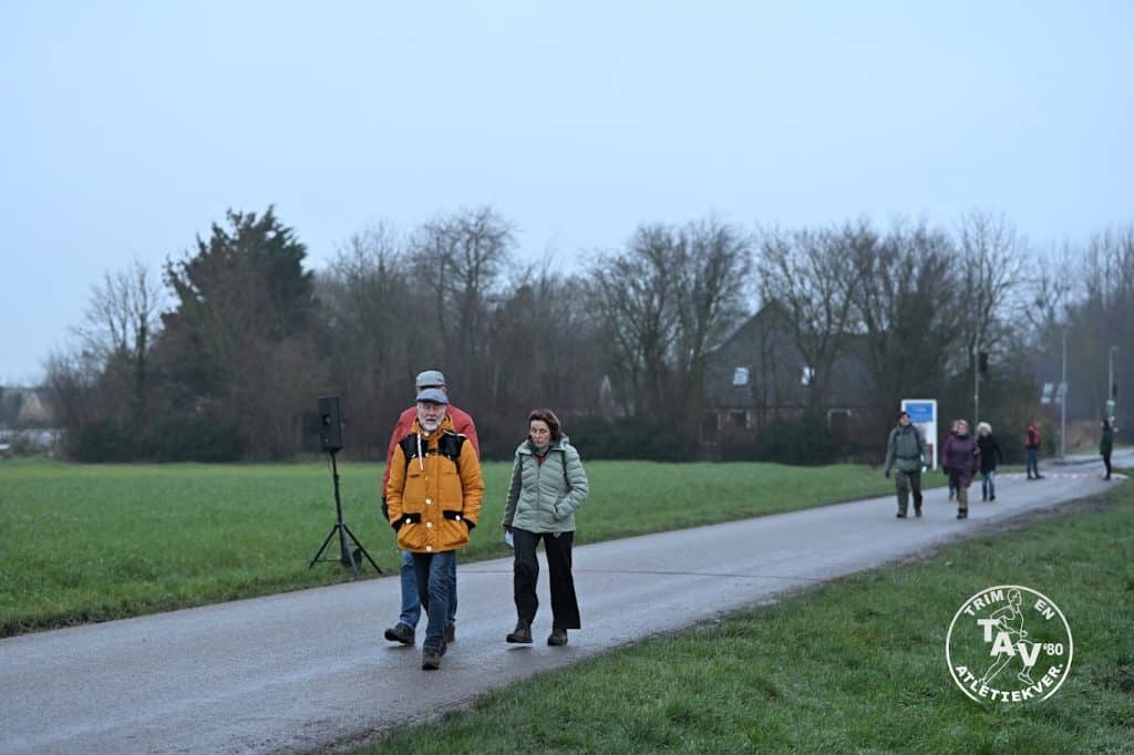Deelnemers aan de wandeltocht van de Snertloop in ’t Veld lopen over een landelijke weg door het buitengebied. Langs de route staan enkele toeschouwers en in de verte zijn bomen en woningen zichtbaar.